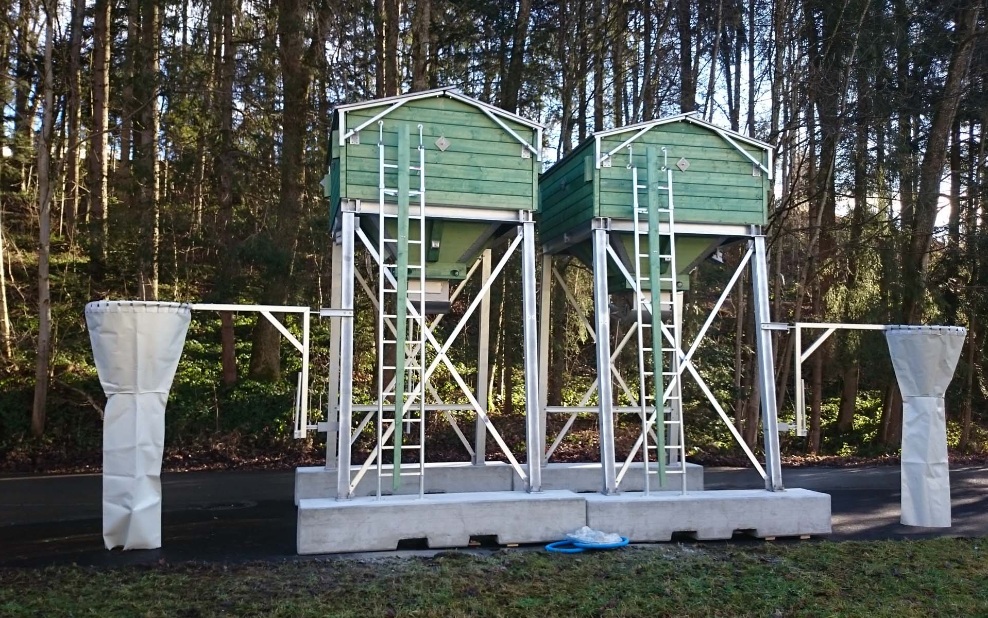 Two small, green, 5 m³ four-sided timber silos on steel substructure, in front of a forest