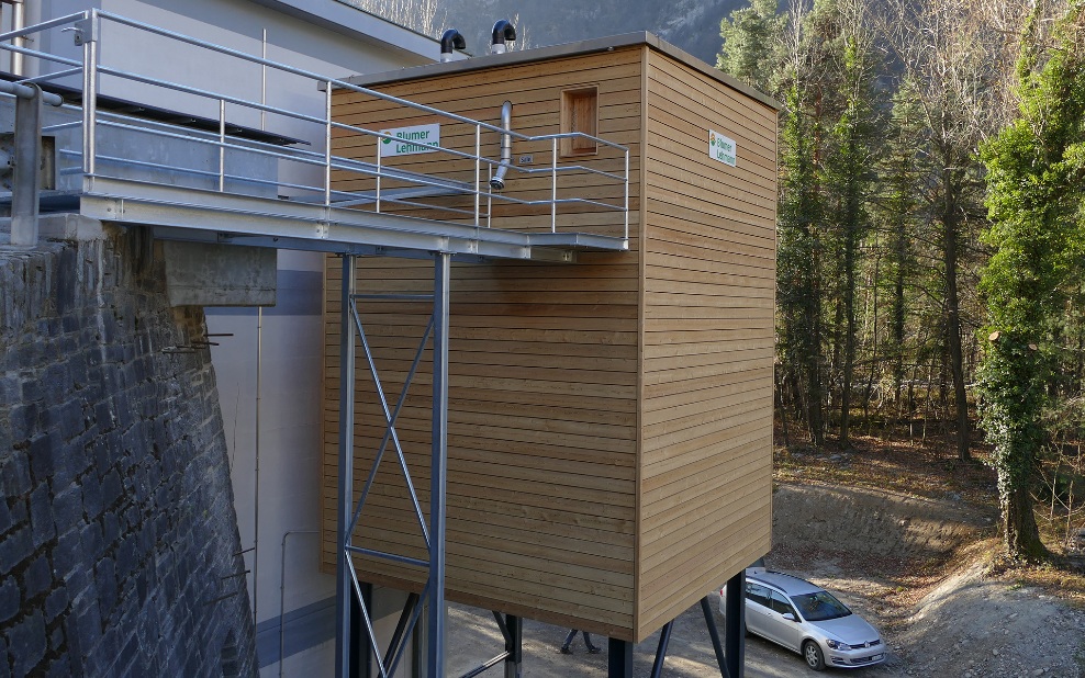 Silo modulaire en bois de 75 m³ avec support en acier gris et accès via une passerelle en acier, accolé à un bâtiment en bois existant