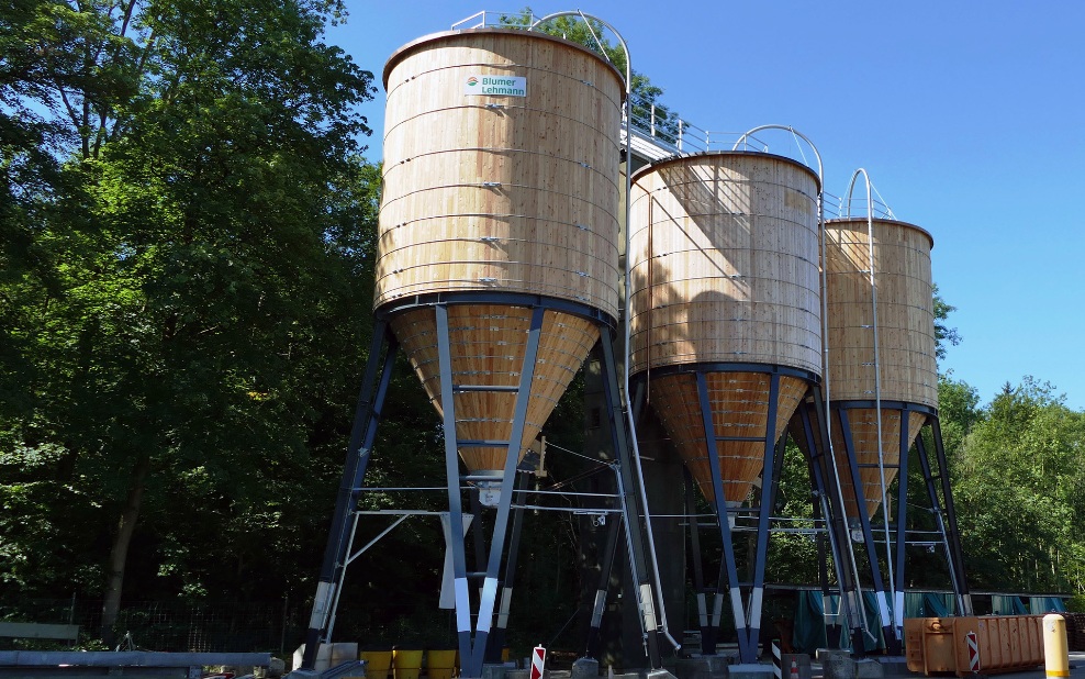 Trois silos ronds en bois de 200 m³ avec support en acier placés côte à côte devant un groupe d’arbres