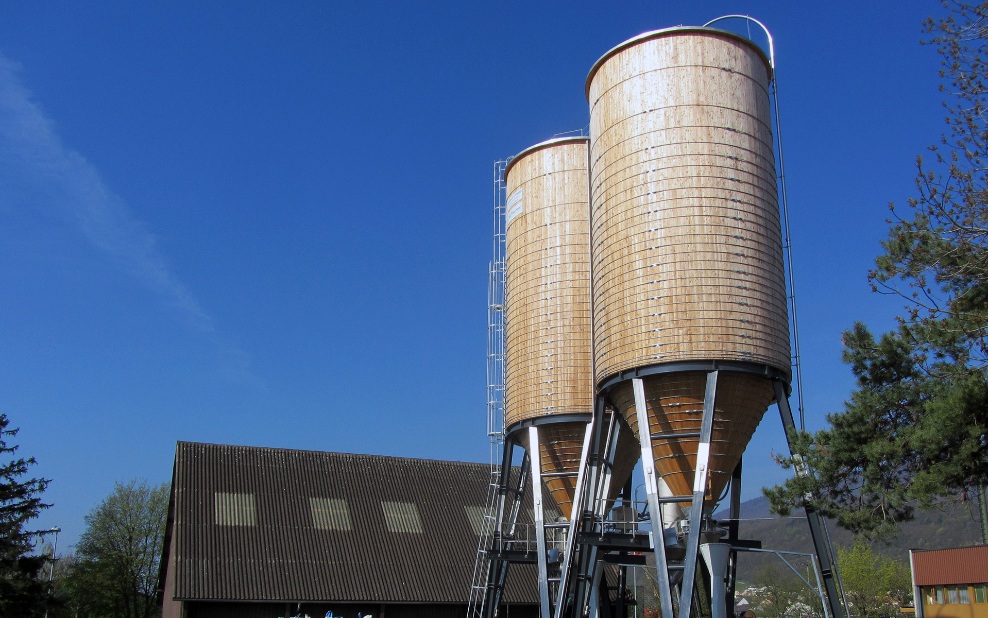 Two 400 m³ round timber silos with steel ladder and grey steel substructure, in front of a barn