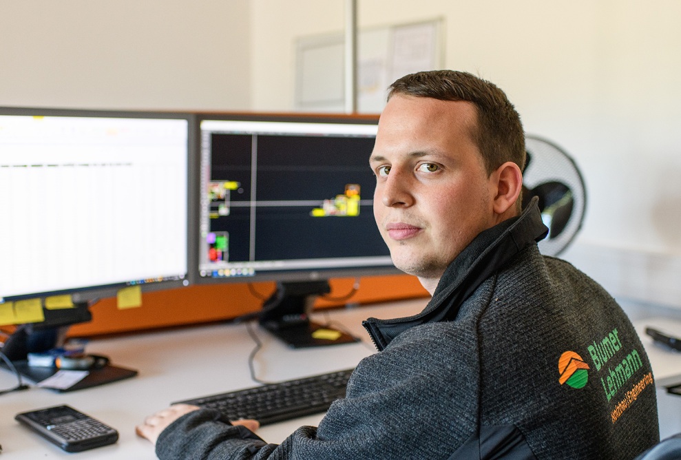 A young man sitting in front of two screens and looking over his shoulder at the camera. He is wearing a jacket with the company logo.