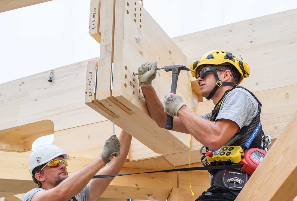 Deux collaborateurs avec casques sont assis sur la construction en bois et fixent des clous.