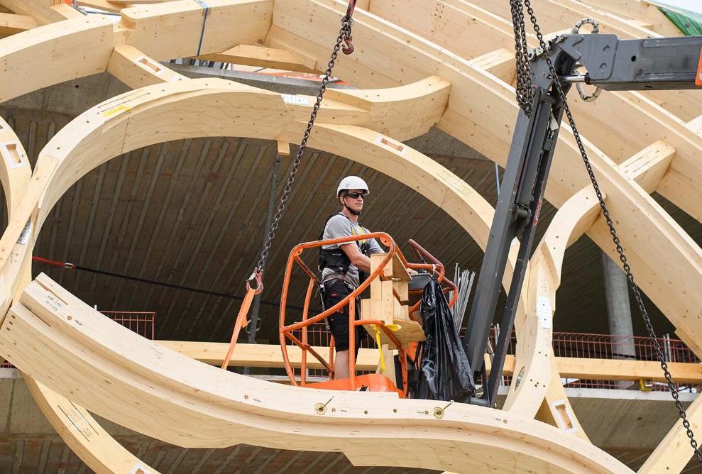 Ein Montagearbeiter befindet sich mit einem orangen Hebekran inmitten der Holzkonstruktion des Swatch-Gebäudes