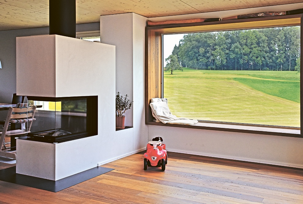 Living and dining area with fireplace in the single-family house bathed in light. View through the large window with wide wooden frame onto a green meadow and nearby forest