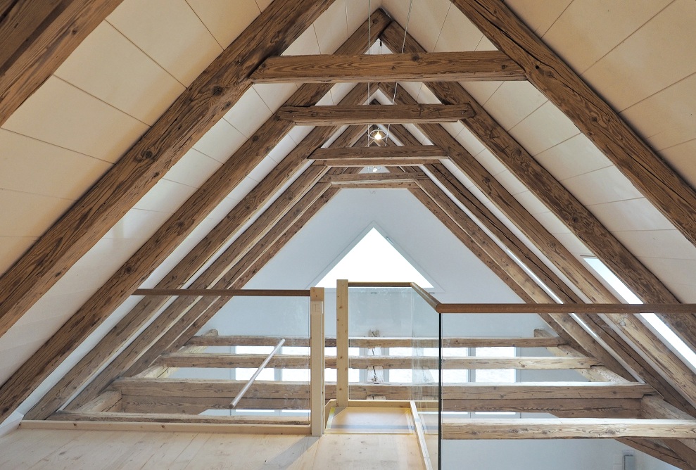 Lichtdurchflutetes offenes Dachgeschoss gesichert durch Glas- und Holzgeländer. Die Dachschrägen sind verkleidet mit breiten Holzbalken.