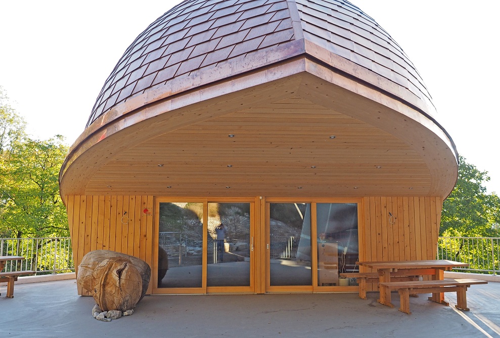 Vue extérieure de la salle d’attente de la station inférieure des remontées mécaniques du Staubern avec deux noix, des bancs et des tables en bois.