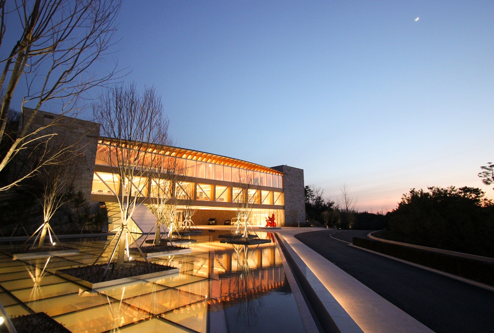 Aussenaufnahme des Haesley Nine Bridges Golfresort Learning Centres. Beleuchtete Holzarchitektur bei Nacht mit Teich im Vordergrund.