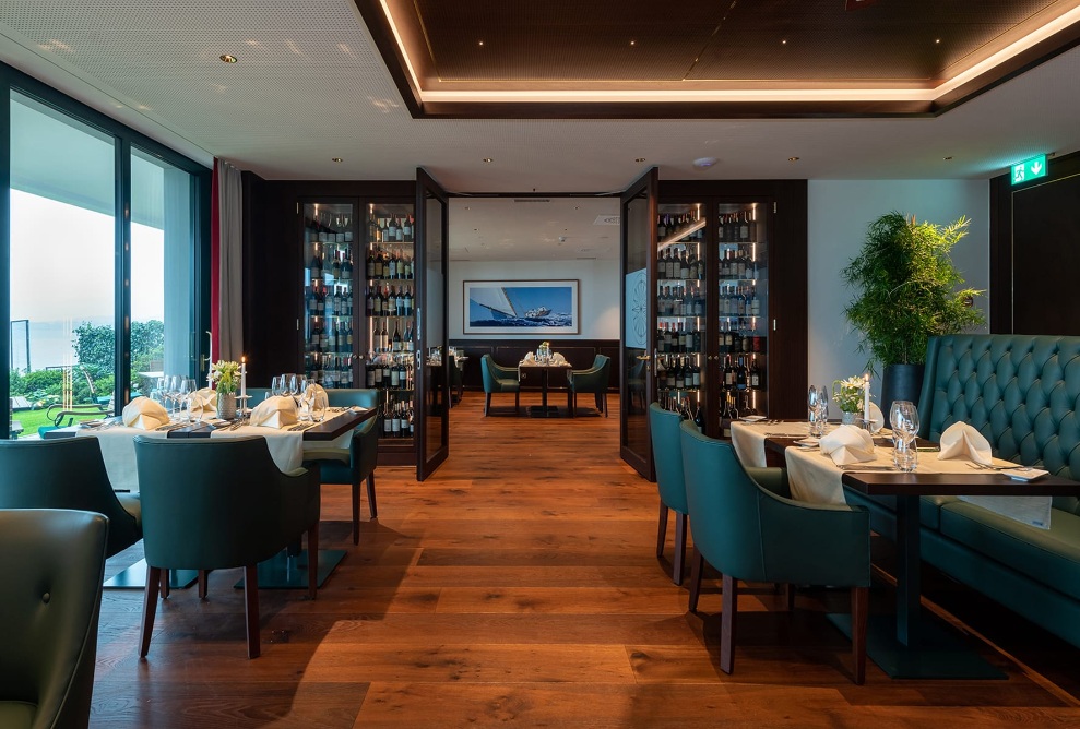 Vue intérieure du restaurant à l'hôtel Bad Horn avec des chaises vert foncé, des tables dressées, un plancher en bois foncé et une grande baie vitrée