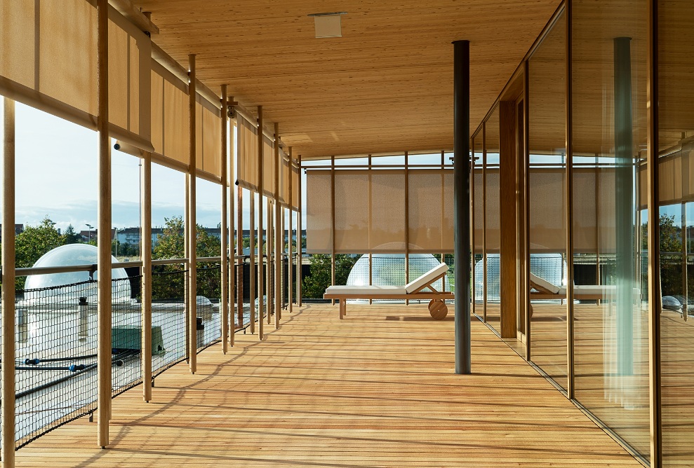 Veranda with a lounger; roller blinds to provide shade on one side and a glazed building facade on the other