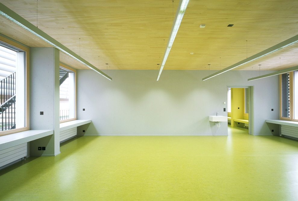 Blick in ein leeres Schulzimmer mit Fensterfronten auf beiden Seiten, gelbem Boden und Fichtendecke. 