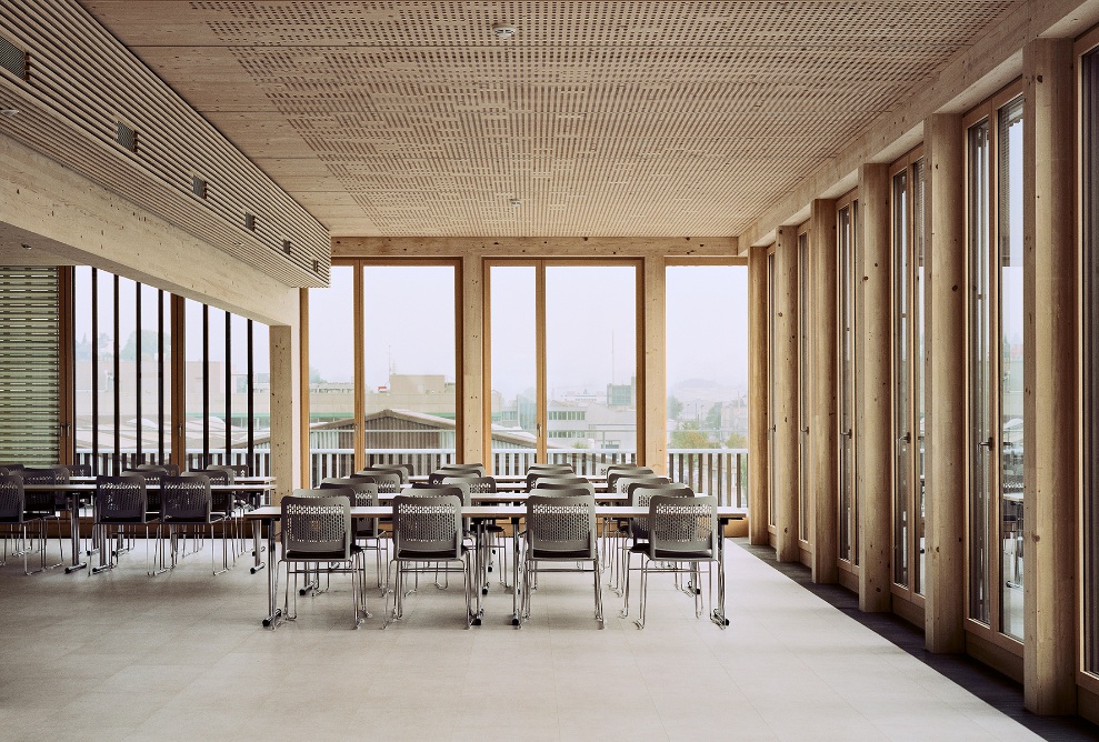 Der Aufenthaltsbereich ist mit langen Tischen und Sitzmöglichkeiten ausgestattet. Der Raum ist durch bodenlange Fenster lichtdurchflutet und die Wände sowie die Dachstruktur sind komplett aus Holz.