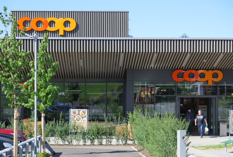 Haupteingang des Coop Super Center in Uzwil mit dunkler Holzfassade sowie Vorplatz, kleinen Bäumen und Kleinpflanzen.