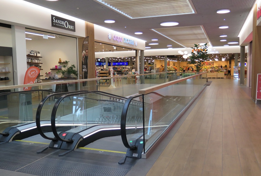 Im Eingangsbereich des Coop Super Centers Uzwil mit den Eingängen zu den Geschäften sowie Rolltreppen und Fussboden aus hellem Holz.