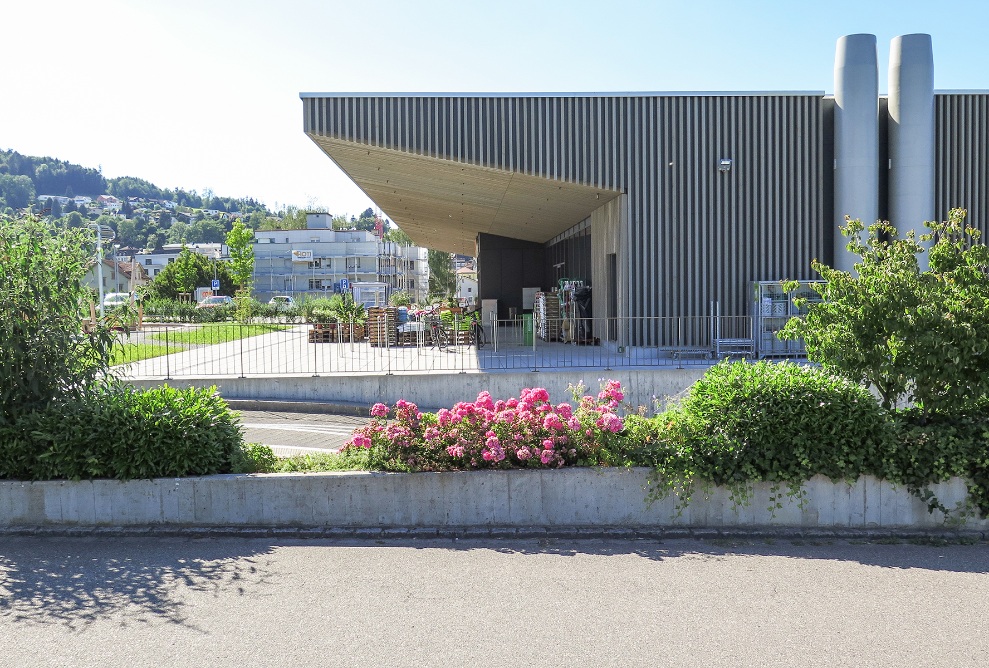 Le Coop Super Center vu de côté avec sa façade en bois prégrisé et l’auvent de la terrasse du restaurant.<br/>