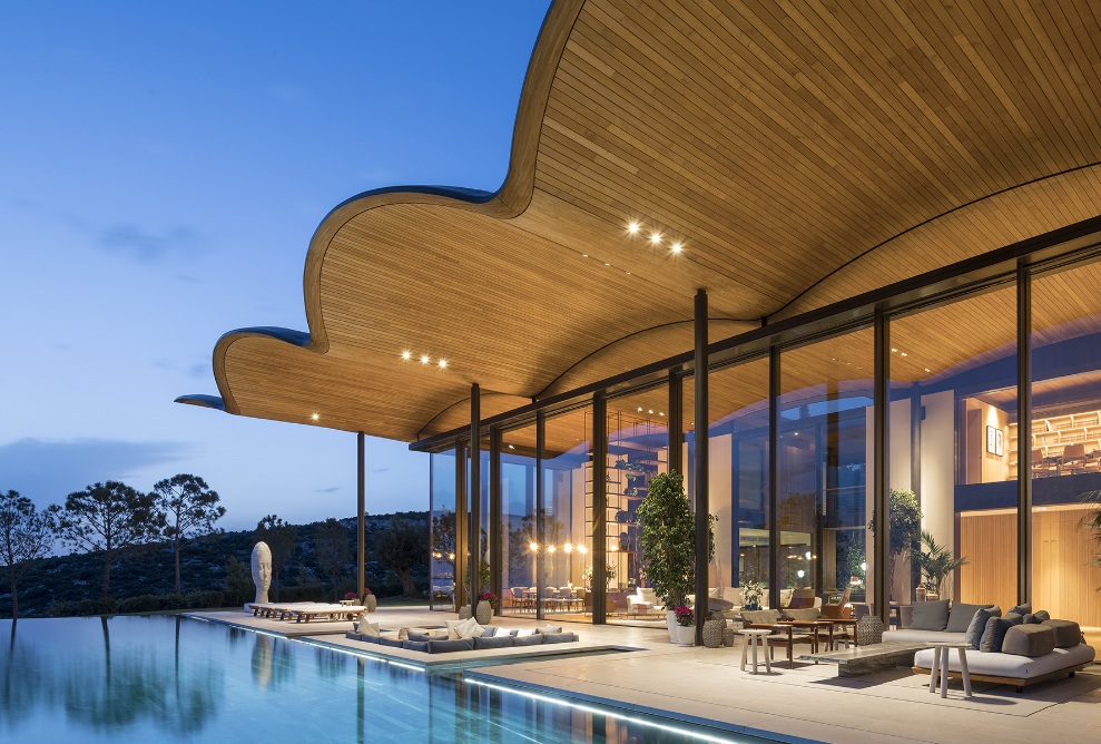 Prise de vue de la piscine d’une villa privée à Bodrum. La zone d’entrée de la villa est entièrement vitrée avec un toit en bois clair.