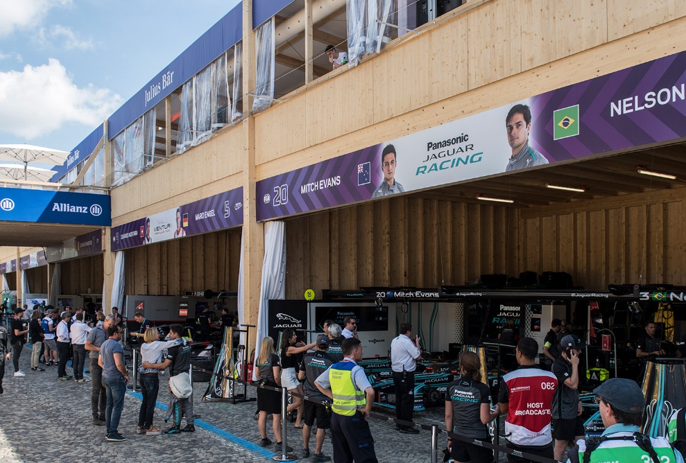 Le bâtiment événementiel en bois a servi de ligne des stands au Zurich E-Prix 2018. Au premier plan, on voit des visiteurs et des membres des équipes. En arrière-plan, les voitures de course sont dans les stands.