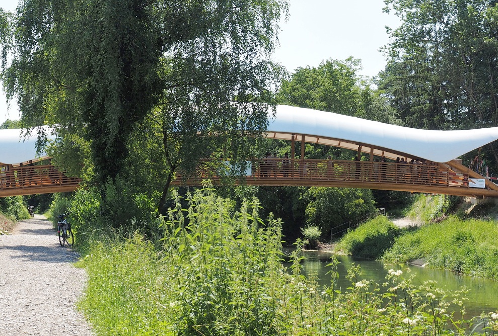 Die Aufnahme zeigt die Holzkunstbrücke etwas versteckt hinter einem grossen Baum bei schönem Wetter