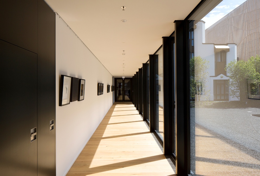 Un couloir d’hôtel baigné de lumière à l’hôtel Säntispark, décoré de tableaux et doté d’un plancher en bois clair. Les baies vitrées rendent le couloir accueillant et lumineux.