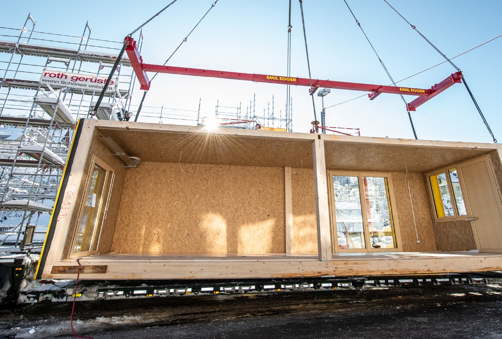 Timber module on a crane: truck is unloaded on the construction site in bright winter sunshine.