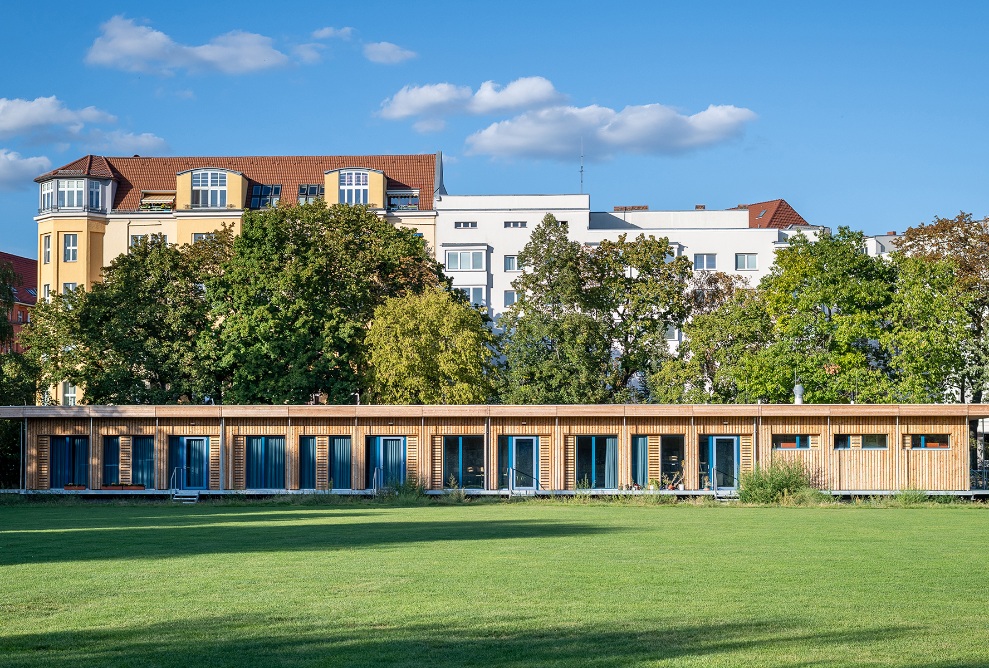 Visualisierung eines eingeschossiges temporären Schulhauses in Berlin Schönefeld. Die Holzfassade hebt sich von der grünen Wiese und vom blauen Himmel ab.