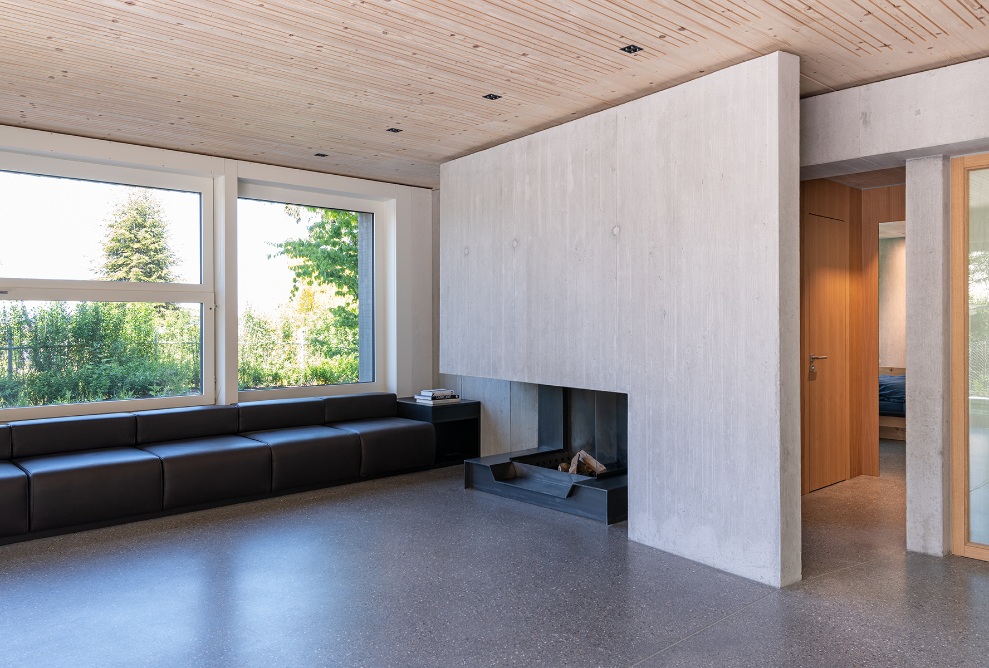 Salle de séjour dans la Sutra House dotée d’une grande baie vitrée, de murs en béton, d’un plafond et d’une porte en bois ainsi que d’une cheminée et d’un canapé. 