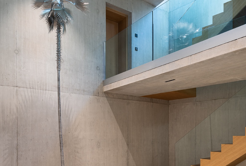 Hoher Raum mit viel Beton, Glas, Holztreppe und einem Kunstwerk in Palmen-Form.