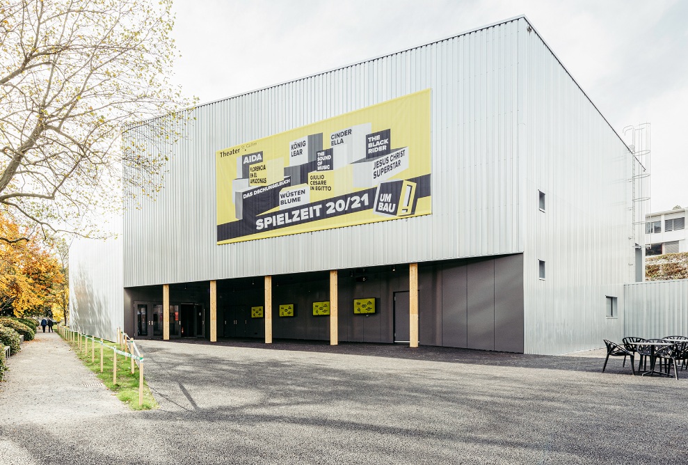 Theaterprovisorium mit hellgrauer Holzfassade und schwarz gestrichenem Eingangsbereich 