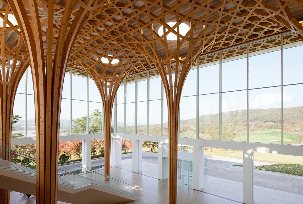 View of the two-storey hall of the golf club. The wooden supporting structure dominates the room while the vast golf course can be seen through the huge window in the background.