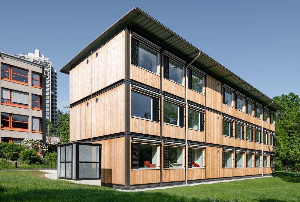 Gesamtansicht des ZM10-Schulpavillons Sihlweid in Zürich auf dem Schulareal und mit Schulhaus im Hintergrund