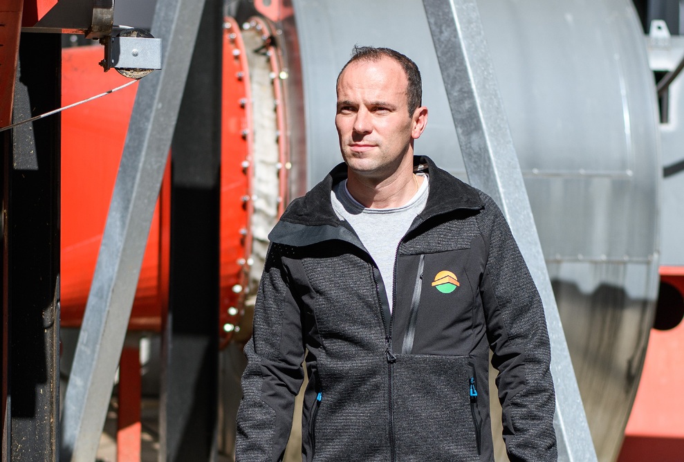 Ein Mann steht vor einem roten Hintergrund. Er trägt eine schwarze Jacke und ein graues Shirt. 