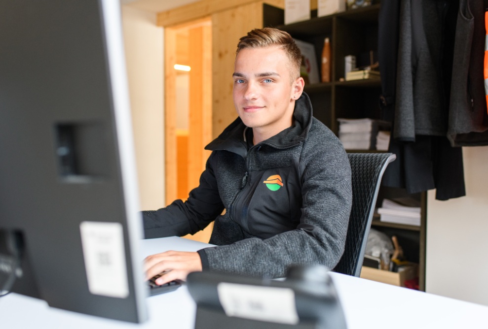 1 Lernender sitzt auf einem Stuhl am Bürotisch und bedient den Computer. Der Lernende schaut in die Kamera. 