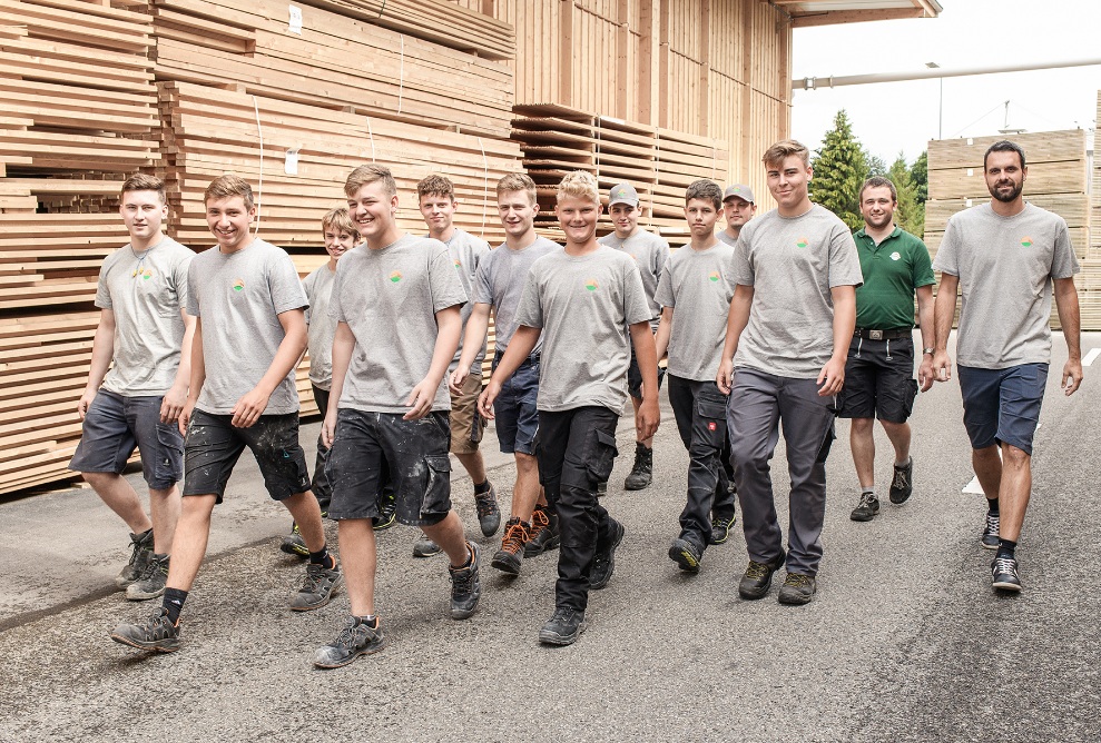 A group picture with eleven young people in training to become carpenters and two trainers walk in front of large stacks of wood and laugh