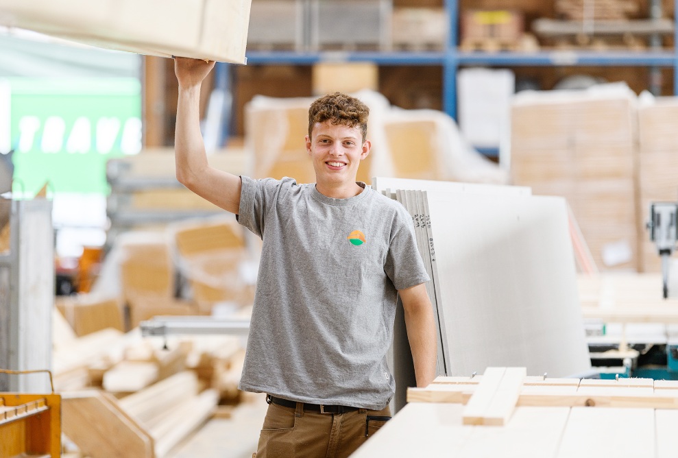 Ein Lernender steht hinter einem Palett Holzbretter in der Produktion und hält sich mit einer Hand an einem Holzkeil fest