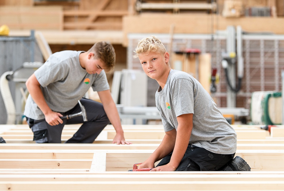 Two apprentice carpenters kneel on a wooden construction, both have tools in their hands and work