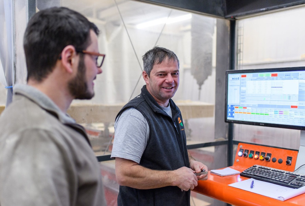 2 Personen sind im Führerstand einer CNC-Maschine zu sehen. Im Hintergrund sind 2 PCs die bedient werden. Eine Person schaut in die Kamera, eine auf den Bildschirm.