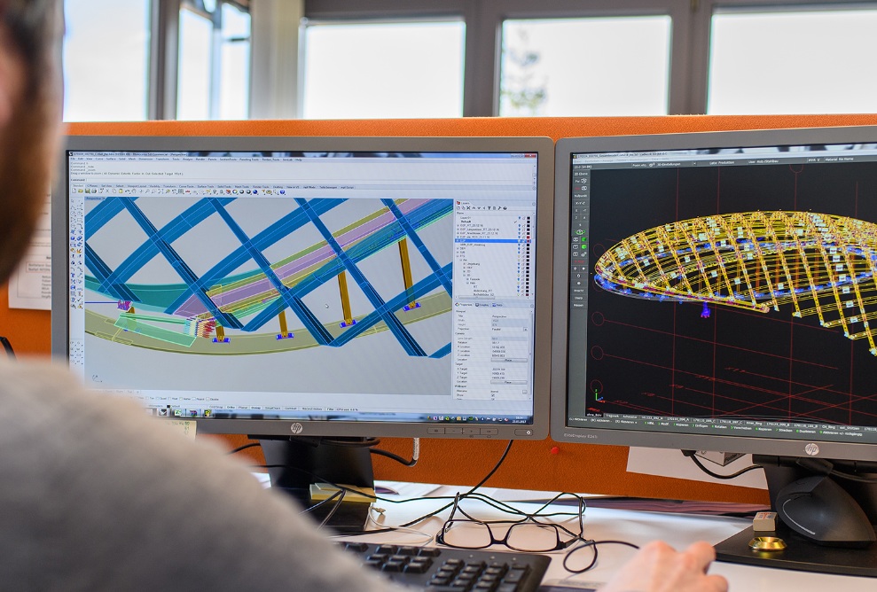 Ein Mitarbeitender sitzt an zwei Bildschirmen und plant am 3D-Modell eines Baus.