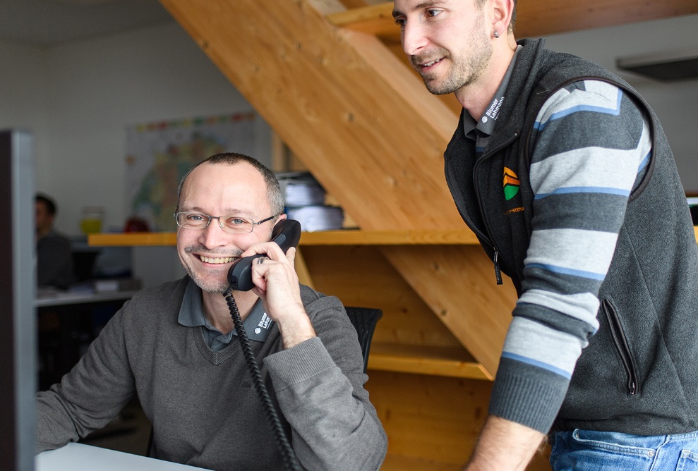 Zwei Mitarbeitende in einer Besprechungssituation wobei einer am Telefon ist und der andere stehen.