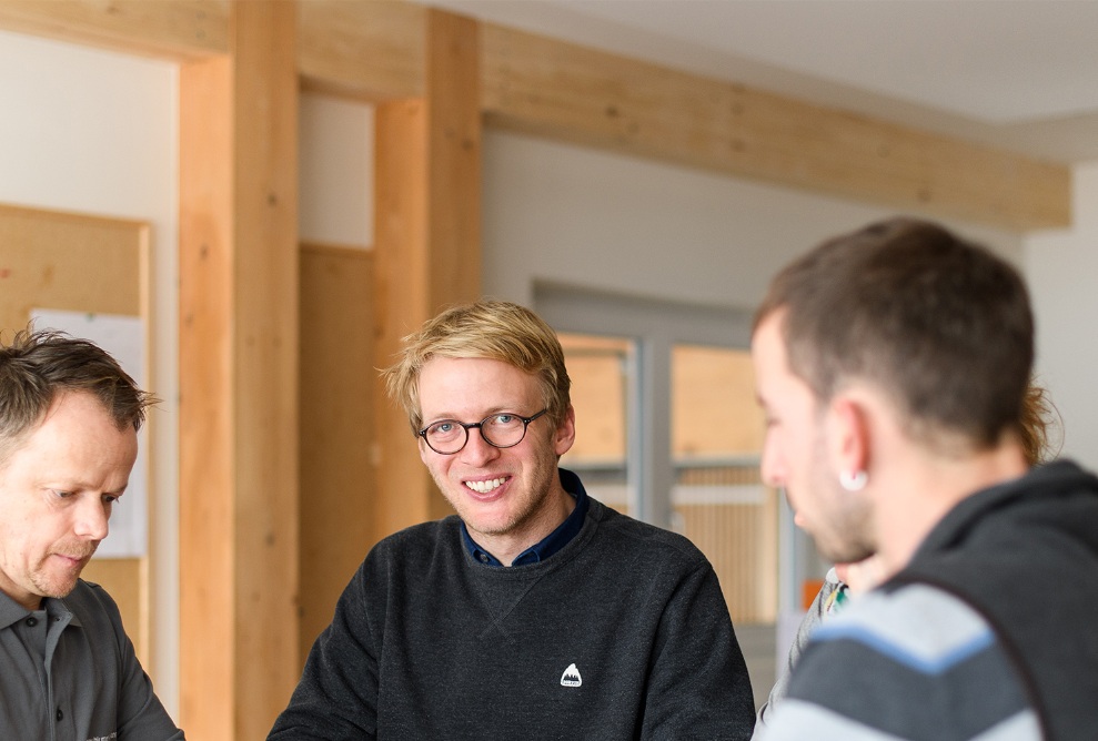Team discussing a plan at a standing desk. The man in the middle is wearing glasses and a black pullover.