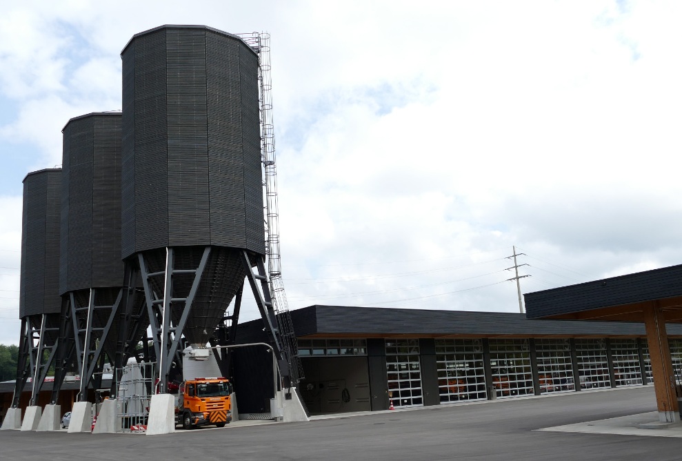Three black, 900 m³, twelve-sided timber silos, next to a modern maintenance depot building with the identical black facade